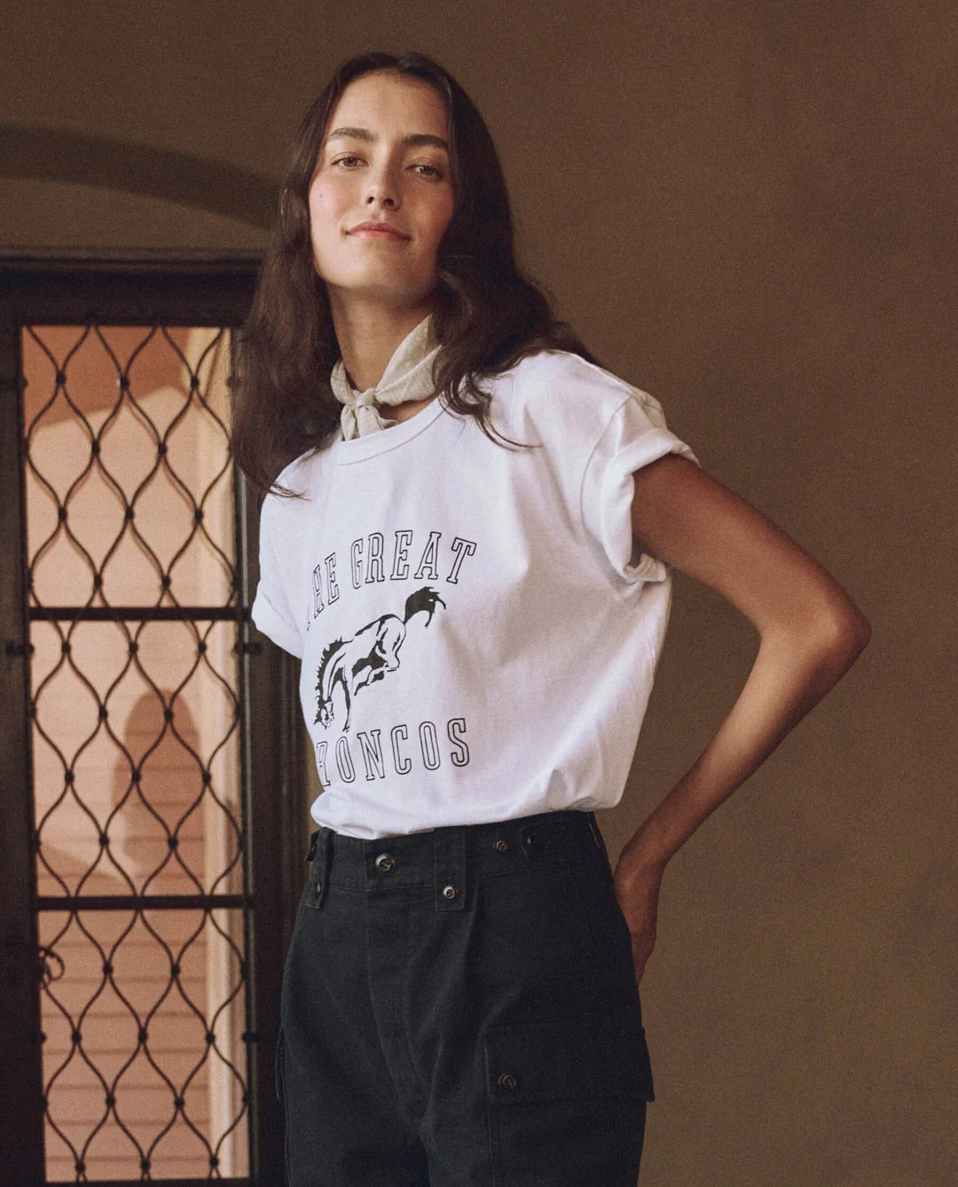 A person with long hair is wearing The Great Inc.'s The Boxy Crew w/ Bucking Bronco tee, featuring "THE GREAT BRONCOS" text and a western bronco graphic. They have a white scarf around their neck, standing indoors before a handcrafted Los Angeles wrought iron door.