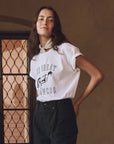 A person with long hair is wearing The Great Inc.'s The Boxy Crew w/ Bucking Bronco tee, featuring "THE GREAT BRONCOS" text and a western bronco graphic. They have a white scarf around their neck, standing indoors before a handcrafted Los Angeles wrought iron door.