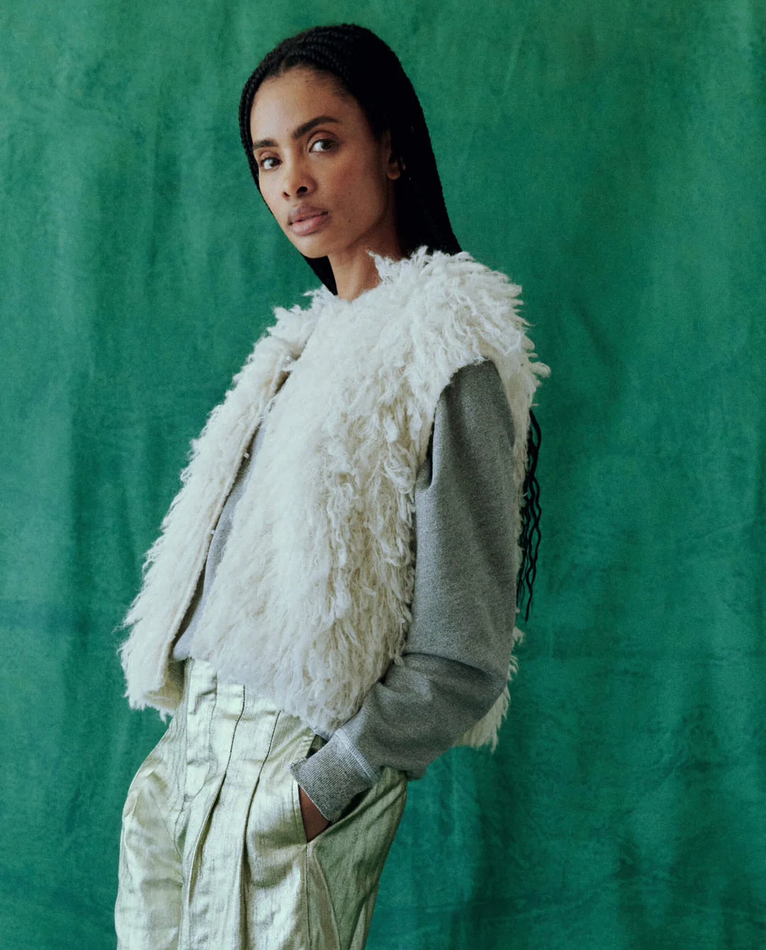 A person with long hair stands against a green textured background, confidently looking at the camera while wearing The Boucle Loop Vest by The Great Inc. over a gray long-sleeve shirt and metallic trousers.