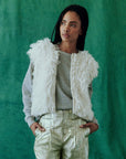 A person stands against a green background, wearing a gray sweater paired with The Great Inc.'s Boucle Loop Vest, crafted from fur alternative. Their metallic gold pants complement their long braided hair as their hands rest casually in their pockets.