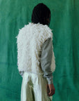 A person with braided hair is standing with their back to the camera, showcasing The Boucle Loop Vest by The Great Inc., featuring a fluffy white fur-alternative design over a gray sweater and metallic pants. The plain green wall serves as the backdrop, accentuating the stylish ensemble.