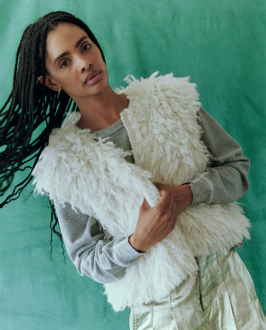 A person with long braided hair stands against a green background, wearing The Great Inc.'s Boucle Loop Vest—a fluffy white piece made from fur alternative alpaca boucle yarn—layered over a gray shirt.