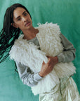 A person with long braided hair stands against a green background, wearing The Great Inc.'s Boucle Loop Vest—a fluffy white piece made from fur alternative alpaca boucle yarn—layered over a gray shirt.