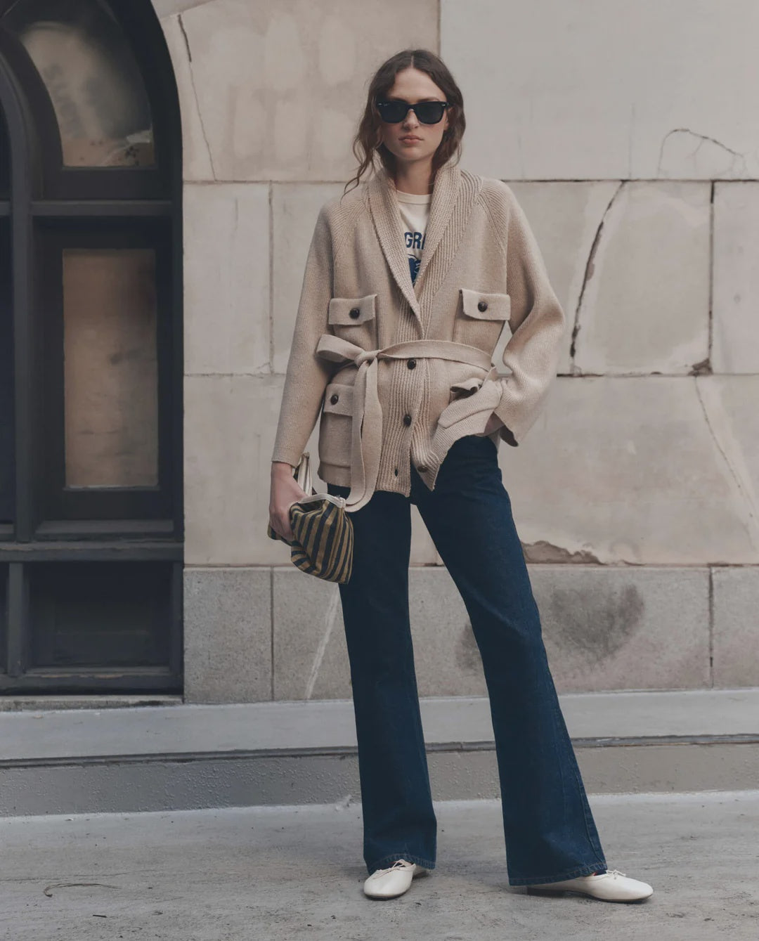 Woman outdoors in wide-leg jeans with striped handbag, styled by The Great Inc.; casual modern setting.