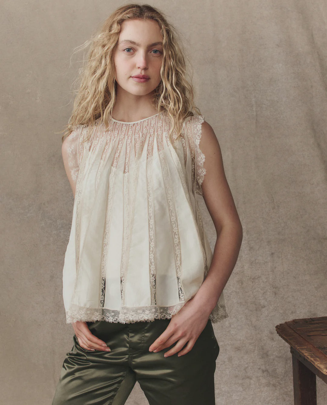 A young woman with wavy blonde hair wears The Keepsake Lace Shell Top by The Great Inc. with olive green pants, standing against a neutral, textured background and facing the camera with a relaxed expression.