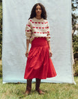 A person with long curly hair stands outdoors against a light backdrop, sporting "The Heart Crew" wool blend sweater by The Great Inc., adorned with red heart motifs, paired perfectly with a red skirt and brown boots. Grass and part of a tree complete the serene background.