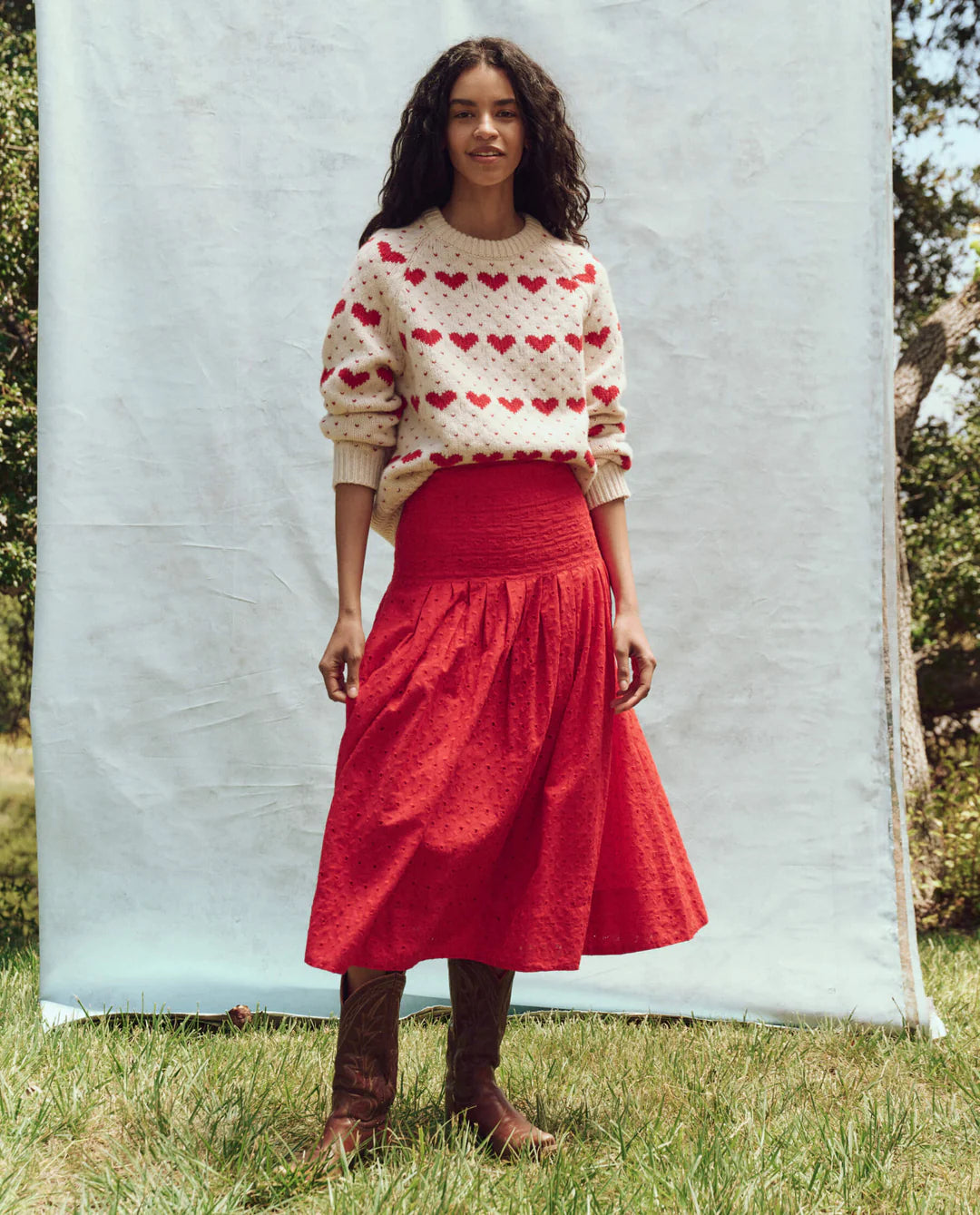 In front of a light backdrop on grass, a person wears The Great Inc.'s cream sweater adorned with red hearts, The Rhythm Skirt in cotton eyelet midi style, and brown boots. Trees fill the background.