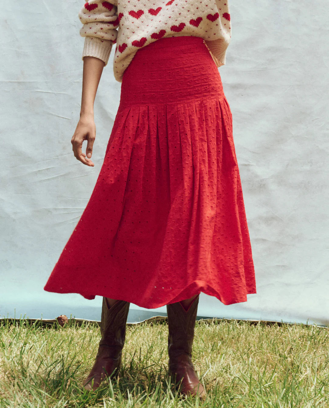 A person stands on grass wearing The Great Inc.'s red midi Rhythm Skirt, cowgirl boots, and a heart-patterned sweater. A pure white fabric backdrop enhances the playful ensemble's rustic charm.