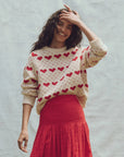 A person with long dark hair smiles against a neutral backdrop, exuding vintage style in The Great Inc.'s cream "The Heart Crew" sweater made from wool and nylon, paired with a red skirt. One hand rests on their head.