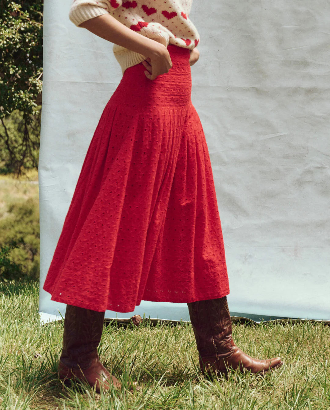 A person stands outdoors, wearing a cream sweater adorned with red hearts, teamed with The Rhythm Skirt by The Great Inc., and brown boots. They are set against lush greenery and a simple white background, evoking the charm of Victorian garments.