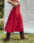 A person stands outdoors, wearing a cream sweater adorned with red hearts, teamed with The Rhythm Skirt by The Great Inc., and brown boots. They are set against lush greenery and a simple white background, evoking the charm of Victorian garments.