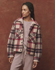 A person with braided hair wears The Smith Jacket by The Great Inc. with a boxy silhouette over a light-colored outfit against a textured brown background. They stand with a relaxed posture, gazing to the side.