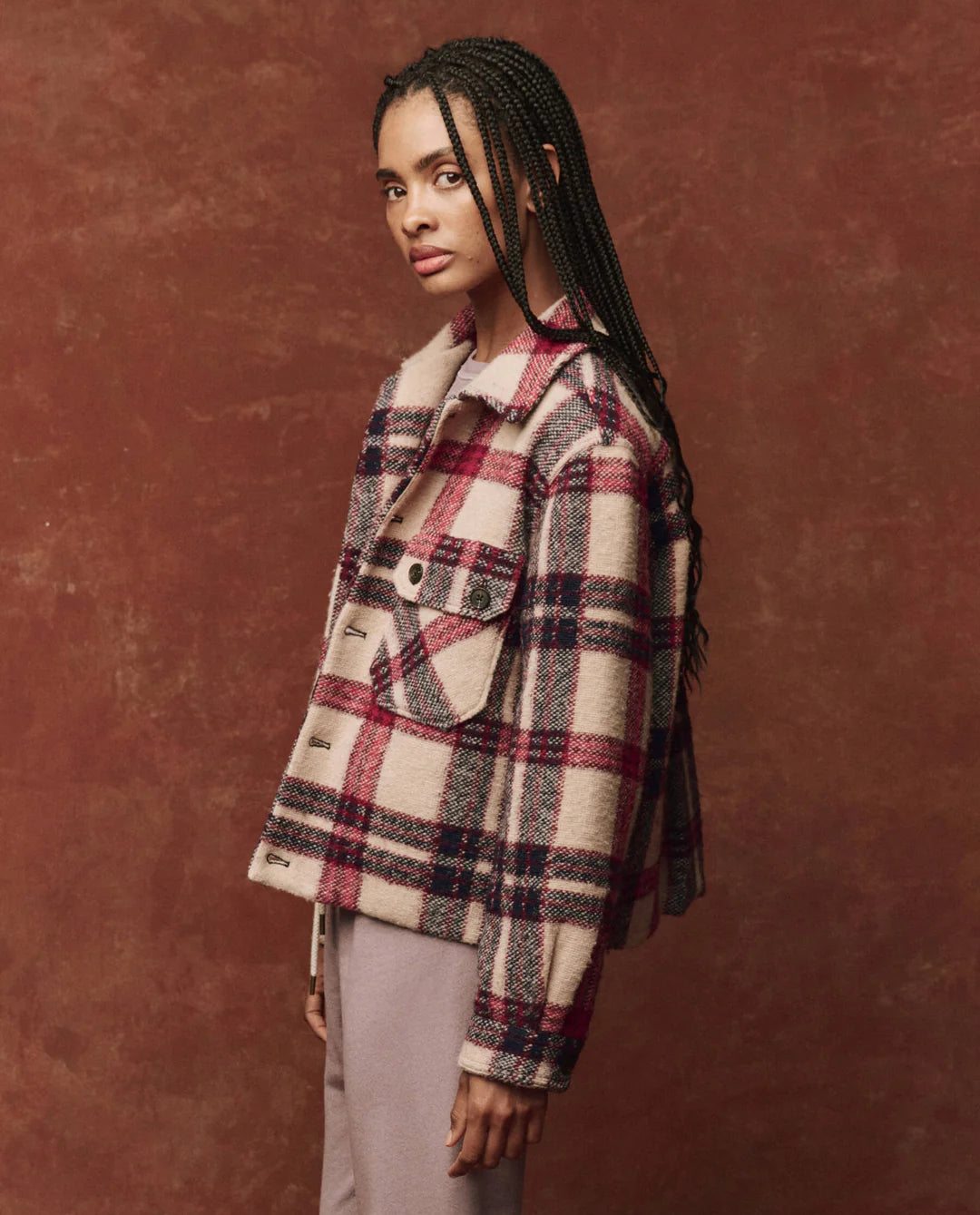 A person with long braided hair is wearing The Smith Jacket by The Great Inc., featuring a vintage logger style in red, white, and black, over a light-colored shirt. They stand against a maroon background, looking to their left.