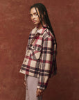 A person with long braided hair is wearing The Smith Jacket by The Great Inc., featuring a vintage logger style in red, white, and black, over a light-colored shirt. They stand against a maroon background, looking to their left.