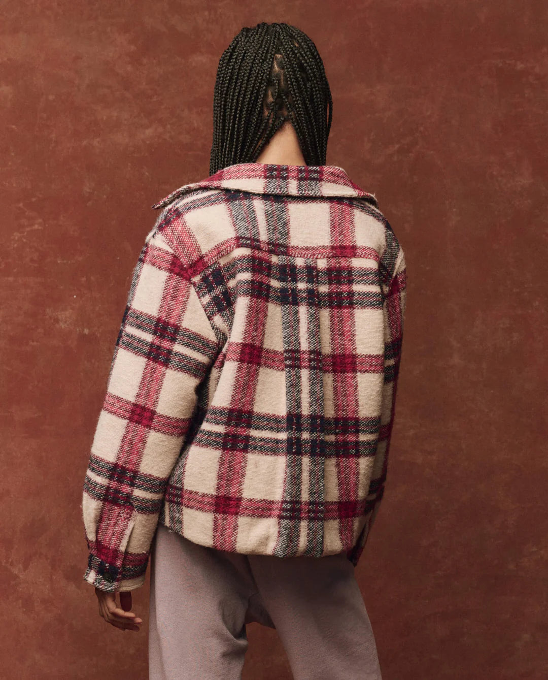 A person with braided hair, wearing The Smith Jacket by The Great Inc., which features vintage colors of red, white, and black with tailored details, stands with their back to the camera against a brown backdrop.