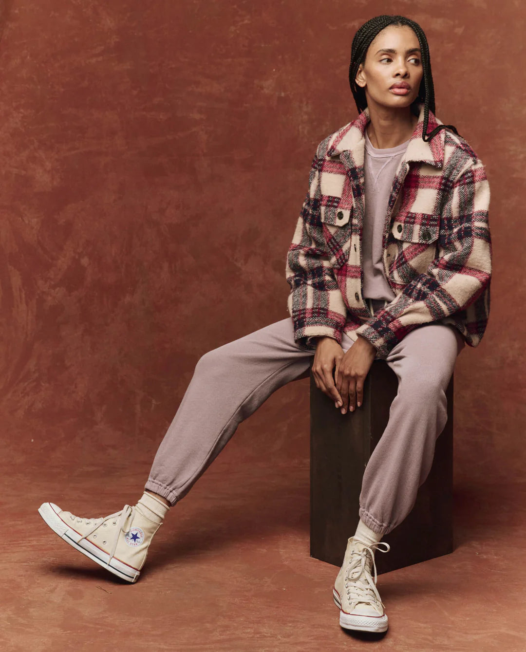 A person with braided hair sits on a block, wearing The Smith Jacket by The Great Inc. over a matching top and pants, paired with high-top sneakers. The rich, textured brown background complements the tailored details of their ensemble.