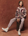 A person with braided hair sits on a block, wearing The Smith Jacket by The Great Inc. over a matching top and pants, paired with high-top sneakers. The rich, textured brown background complements the tailored details of their ensemble.