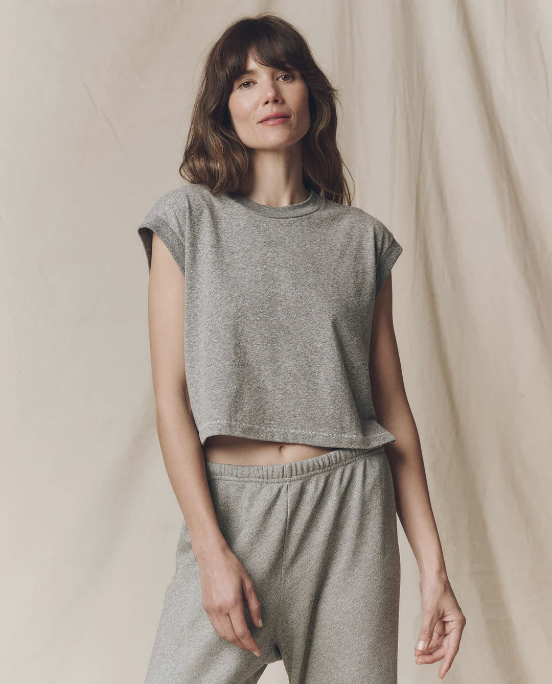 A woman with shoulder-length brown hair wears The Square Tee by The Great Inc., a light gray, sleeveless top, paired with matching sweatpants. She stands before a beige fabric backdrop, gazing past the camera with a relaxed expression.