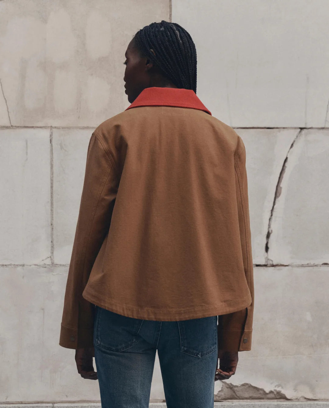 The Great Inc. Swingy Jacket in a casual style, shown with blue jeans against a cracked wall backdrop.