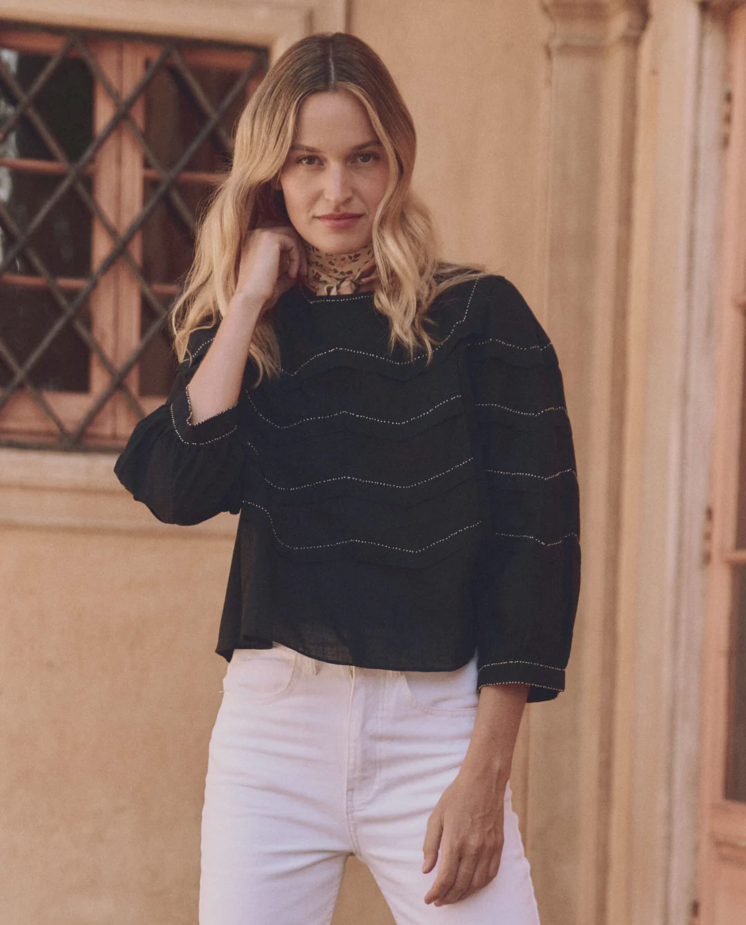 A person with long, blonde hair wears The Great Inc.'s chic Beaded Chevron Top and white pants, standing in front of a building with a beige facade and pleated windows. The person gazes at the camera with a neutral expression.