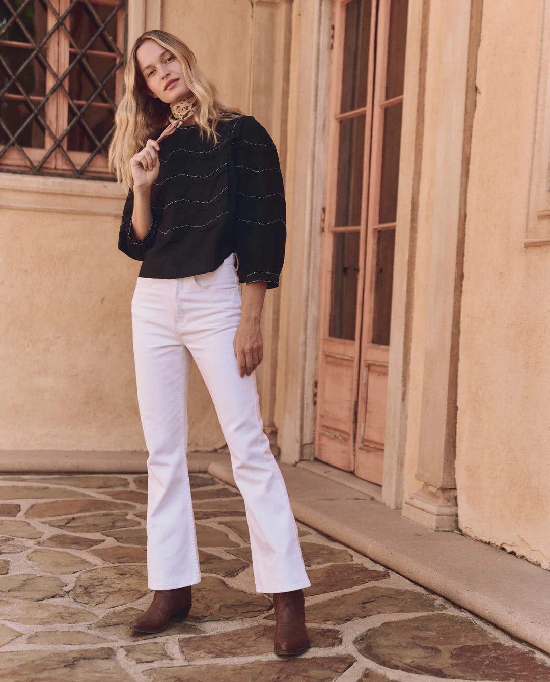 On a stone patio in front of a beige building with a wooden door and window, a person with long hair wears The Great Inc.'s Beaded Chevron Top, white flared pants, brown boots, and a patterned scarf showcasing vintage-inspired beadwork.