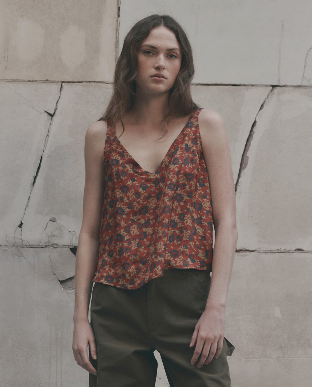 The Port Cami by The Great Inc. in white, styled with olive pants against a textured concrete wall backdrop.