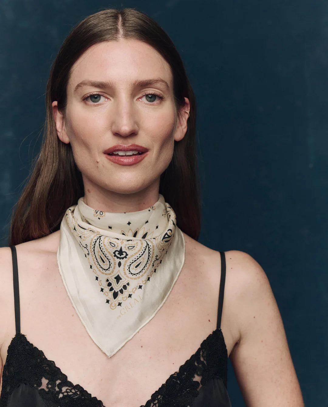 A woman with straight brown hair wears The Great Inc.'s The Silk Bandana in paisley around her neck and a black lace camisole, standing against a dark blue background and looking forward with a slight smile.