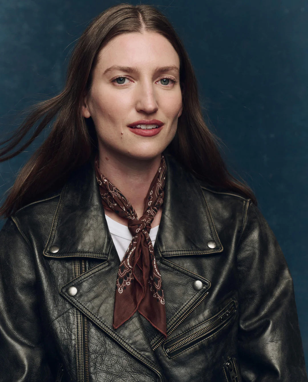 A woman with long brown hair, wearing a black leather jacket over a white shirt and The Silk Bandana by The Great Inc. as a brown patterned scarf, poses against a dark blue background, looking at the camera with a slight smile.