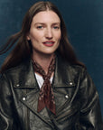 A woman with long brown hair, wearing a black leather jacket over a white shirt and The Silk Bandana by The Great Inc. as a brown patterned scarf, poses against a dark blue background, looking at the camera with a slight smile.