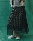 A person stands against a teal backdrop wearing The Era Skirt by The Great Inc., featuring embroidered lace over a beige layer, paired with a black and white striped sweater. Floral patterned socks peek from their beige sneakers. The image is cropped at the shoulders, capturing this chic ensemble.