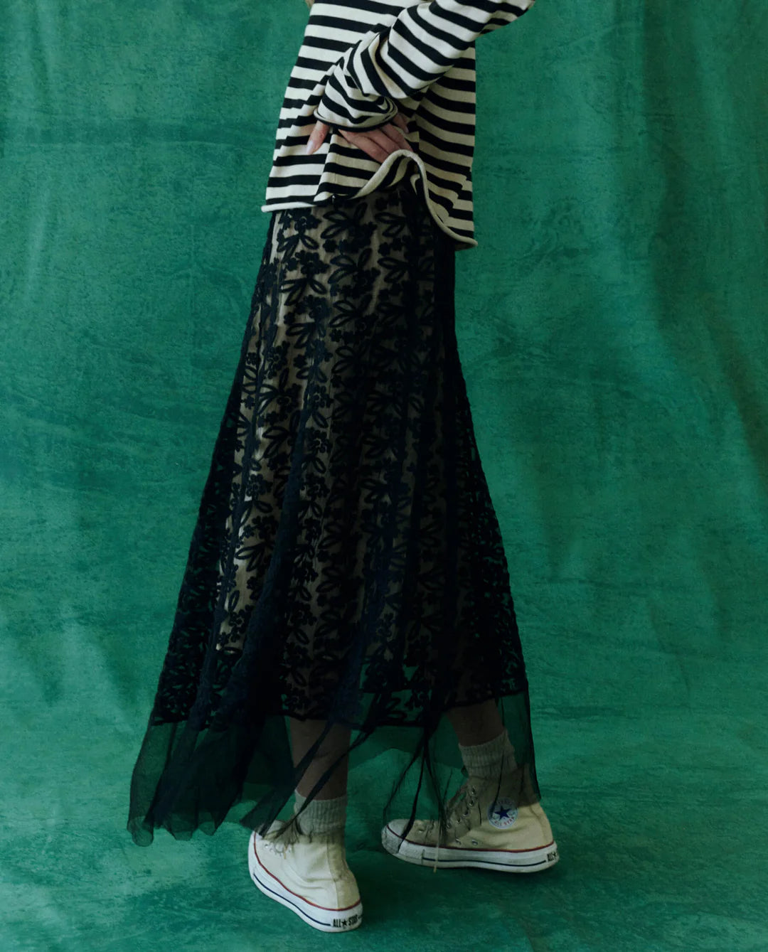 A person stands against a green background, wearing The Era Skirt by The Great Inc., a vintage-inspired black embroidered lace piece layered over white pants. They pair it with white sneakers and a black-and-white striped top, hands resting on their waist, though their head is not visible in the image.