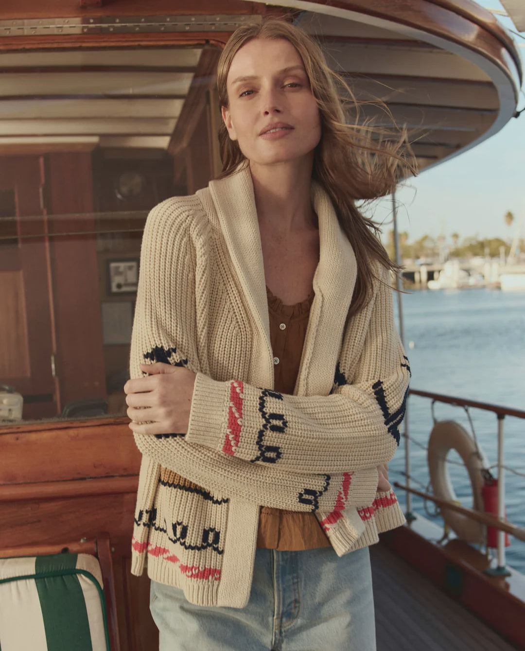 A woman with long hair stands on a boat, wearing The Great Inc. Sailboat Lodge Cardigan—a cotton blend with hand-embroidered boats and classic red and navy patterns—over a brown top and light blue jeans, with a marina in the background.