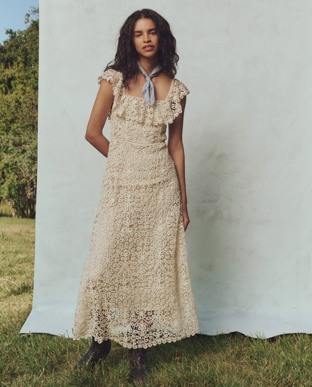 A woman with long, dark hair stands outdoors on the grass, wearing The Balcony Dress by The Great Inc. Its vintage lace design features a fitted bodice. She has a light blue scarf around her neck. A light blue screen and partially visible trees complete the serene scene.