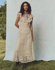 A woman with long, dark hair stands outdoors on the grass, wearing The Balcony Dress by The Great Inc. Its vintage lace design features a fitted bodice. She has a light blue scarf around her neck. A light blue screen and partially visible trees complete the serene scene.