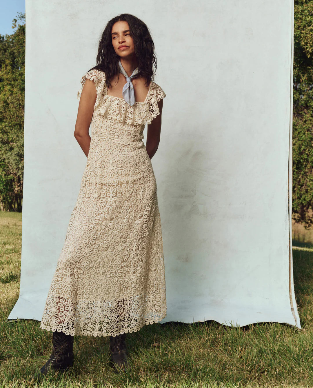 A person stands outdoors on grass in front of a plain backdrop, wearing The Great Inc.'s The Balcony Dress, a long cream lace dress with scalloped edges and a neck scarf. Their hair is styled down, exuding 1930s vintage charm. The sunny ambiance is enriched by trees in the background.