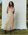 A person stands outdoors on grass in front of a plain backdrop, wearing The Great Inc.'s The Balcony Dress, a long cream lace dress with scalloped edges and a neck scarf. Their hair is styled down, exuding 1930s vintage charm. The sunny ambiance is enriched by trees in the background.