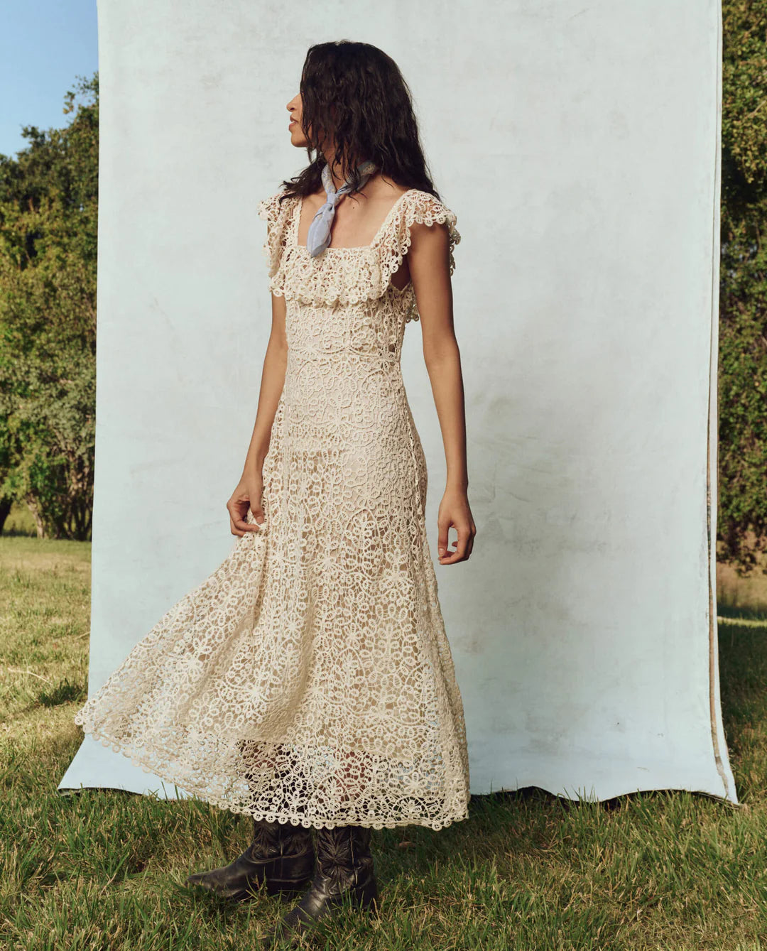 A woman in a long, 1930s vintage The Balcony Dress by The Great Inc. stands outdoors against a light blue backdrop. With long wavy hair and a scarf around her neck, she stands on grass with trees behind her, highlighting the delicate scalloped lace edges of her elegant attire.