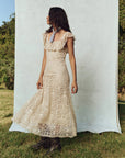 A woman in a long, 1930s vintage The Balcony Dress by The Great Inc. stands outdoors against a light blue backdrop. With long wavy hair and a scarf around her neck, she stands on grass with trees behind her, highlighting the delicate scalloped lace edges of her elegant attire.