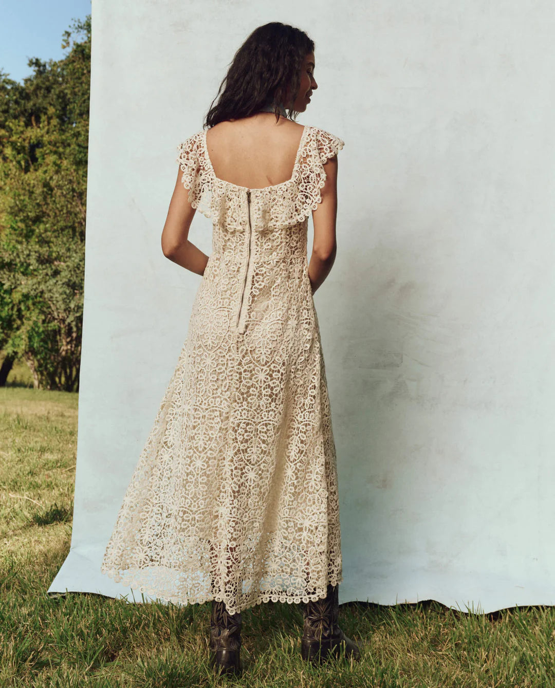 A woman with long hair stands on grass wearing The Balcony Dress by The Great Inc., a sleeveless 1930s vintage lace dress with scalloped edges. Her back faces the camera, and the tranquil setting of trees adds an aura of timeless elegance.