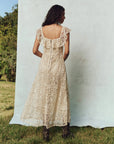 A woman with long hair stands on grass wearing The Balcony Dress by The Great Inc., a sleeveless 1930s vintage lace dress with scalloped edges. Her back faces the camera, and the tranquil setting of trees adds an aura of timeless elegance.