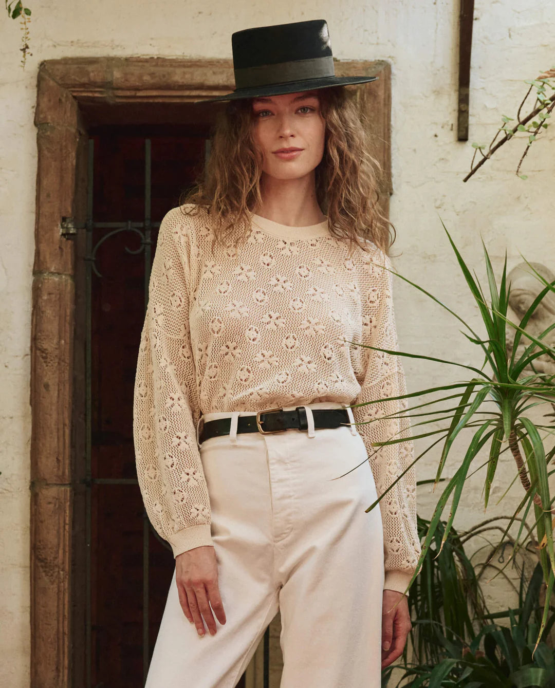 A woman with wavy hair wears The Crochet Pullover by The Great Inc. and stands indoors near a doorway and green plants, pairing it with a black hat, cream high-waisted pants, and a black belt.