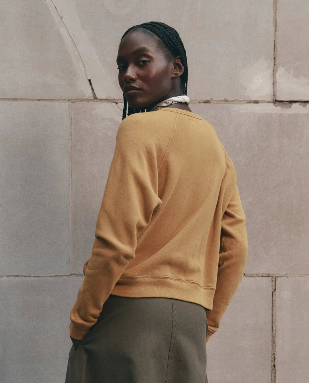 Tan fleece top and olive skirt by The Great Inc. styled in a casual outdoor setting near a stone wall.