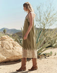 A woman wears The Great Inc.’s The Legacy Dress—vintage-inspired silk with embroidered mesh lace—and cowboy boots, standing outdoors among desert rocks, agave plants, and distant mountains under a clear sky.
