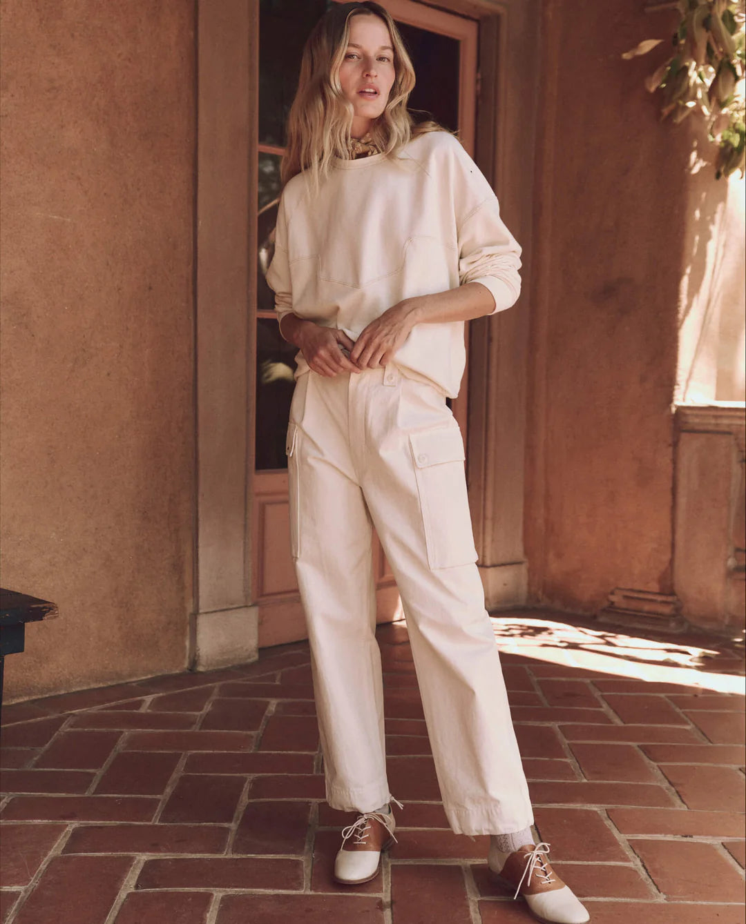 A person stands on a handcrafted terracotta-tiled floor in Los Angeles, wearing The Great Inc.'s cream-colored Western Slouch Sweatshirt paired with matching high-waisted pants with pockets. White shoes with brown accents complete the look against a stucco wall and wooden door backdrop.