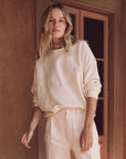 A person with long blonde hair is wearing The Great Inc.'s Western Slouch Sweatshirt and high-waisted pants, standing by a brown wall and glass-paneled door. The casual look features neutral tones reminiscent of relaxed Western wear.