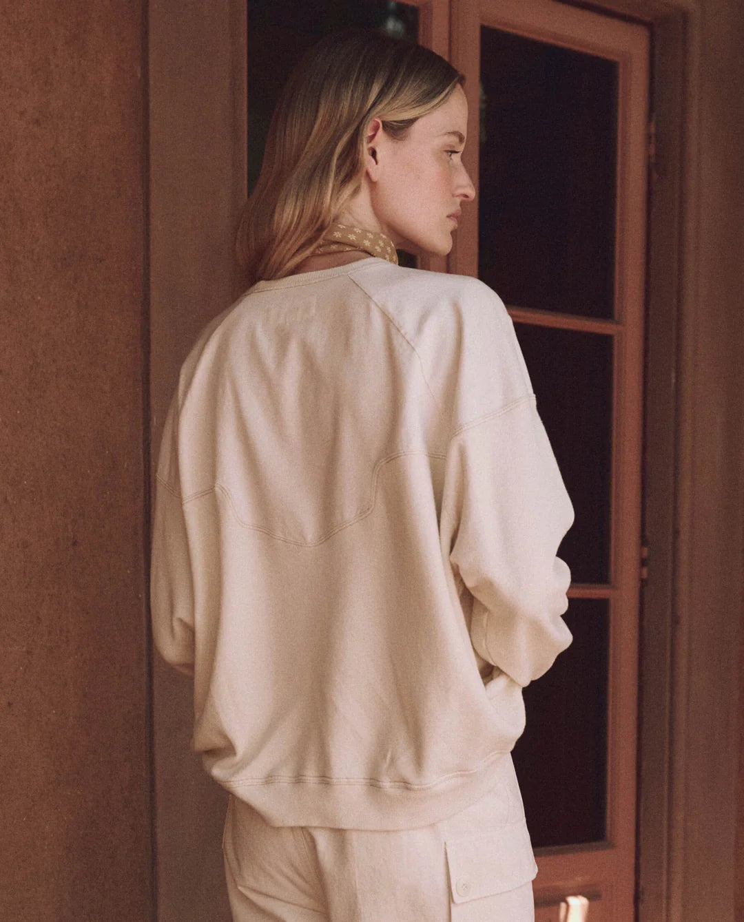 A person with long hair stands facing away, wearing The Great Inc.'s handcrafted The Western Slouch Sweatshirt in off-white. They're near a wooden door, head slightly turned, creating a warm and softly lit ambiance.