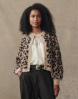A woman with curly hair wears The Windsor Cardigan by The Great Inc.—a cropped leopard jacquard piece in an alpaca blend—over a cream blouse and dark pinstripe trousers, standing against a neutral textured background.