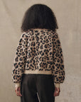 A person with curly hair, seen from behind, wears The Windsor Cardigan by The Great Inc.—a beige and black leopard jacquard alpaca blend sweater—paired with dark pants, standing against a neutral background.