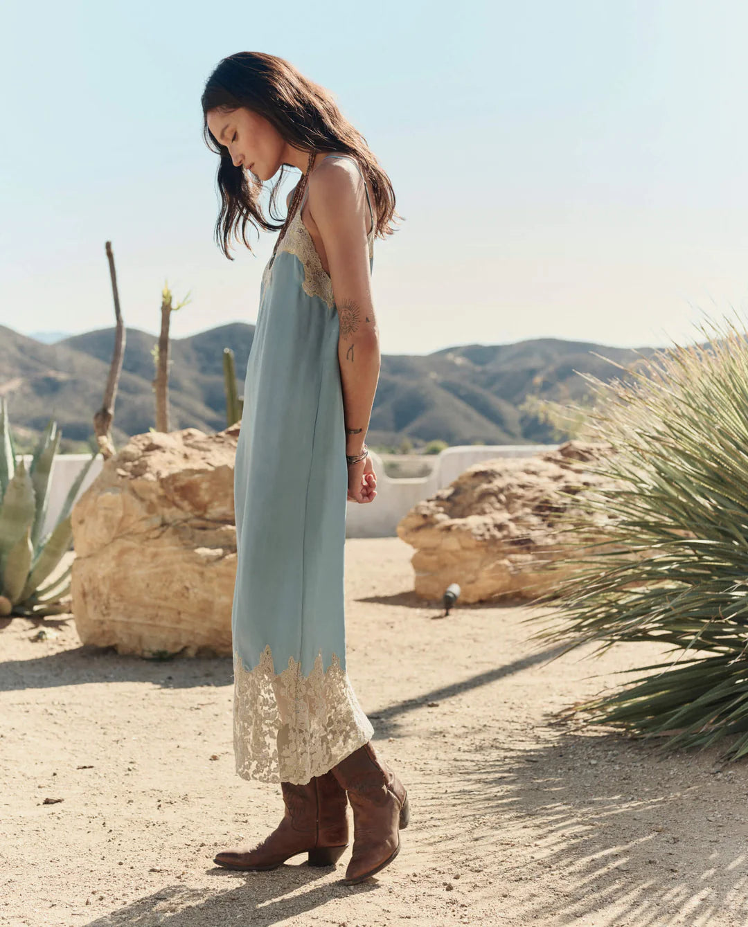 A woman in The Legacy Dress by The Great Inc.—a vintage-inspired piece with embroidered mesh lace trim—stands sideways in a desert landscape amid rocky terrain, cacti, and mountains beneath a clear sky, wearing brown boots.