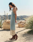 A woman in The Legacy Dress by The Great Inc.—a vintage-inspired piece with embroidered mesh lace trim—stands sideways in a desert landscape amid rocky terrain, cacti, and mountains beneath a clear sky, wearing brown boots.